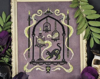 May include: A framed cross-stitch featuring a black cat, cages, and swirling green accents on a lavender background. The artwork is framed in a cream-colored ornate frame. Decorative skulls and foliage surround the framed art.