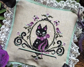 May include: A square, beige cross-stitch pillow with a black cat design in a decorative frame. The cat has a purple bow and is surrounded by purple flowers and green accents. The pillow has a white lace trim and a mint green border.