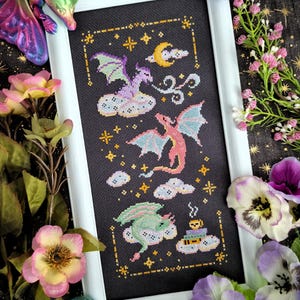 Może przedstawiać: Sztuka haftu krzyżykowego przedstawiająca trzy kolorowe smoki na chmurach, na czarnym tle z gwiazdami i księżycem w kształcie półksiężyca. Oprawiona grafika ma białą obwódkę i jest otoczona kwiatami i kryształami.