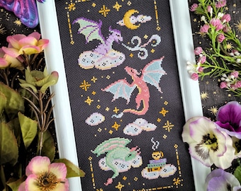 May include: Cross-stitch art featuring three colorful dragons on clouds, set against a black background with stars and a crescent moon. The framed artwork has a white border and is surrounded by flowers and crystals.