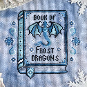 May include: A cross-stitch embroidery on light blue fabric featuring a book titled Book of Frost Dragons with a central blue dragon illustration, surrounded by small blue snowflake motifs and decorative borders in shades of blue and black.