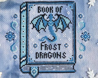 May include: Cross-stitch design featuring a book titled "Book of Frost Dragons" with a blue dragon illustration. The design is in shades of blue and white, surrounded by snowflake and pinecone decorations.