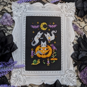 May include: A white frame with a black and white cross-stitch design featuring three cats and a pumpkin. The design includes a crescent moon, stars, and bats.