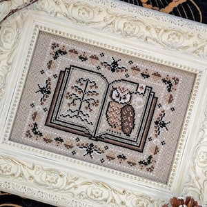 May include: A framed cross-stitch featuring an open book with an owl and tree design. The cross-stitch is in shades of brown and beige, set within a cream-colored, ornate frame. The frame has a detailed floral pattern.