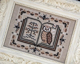 May include: A framed cross-stitch featuring an open book with an owl and tree design. The cross-stitch is in shades of brown and beige, set within a cream-colored, ornate frame. The frame has a detailed floral pattern.
