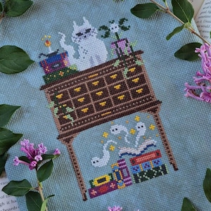May include: Cross-stitch design featuring a brown dresser with a white cat wearing sunglasses on top. The dresser is surrounded by books and ghosts. The background is a light blue fabric, with purple flowers and green leaves.