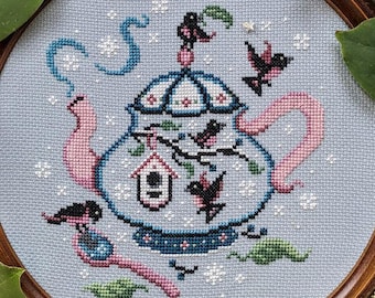 May include: A cross-stitch embroidery featuring a blue teapot with pink accents, surrounded by black birds and a birdhouse. The artwork is framed in a round wooden frame, with white flowers and green leaves around the frame.