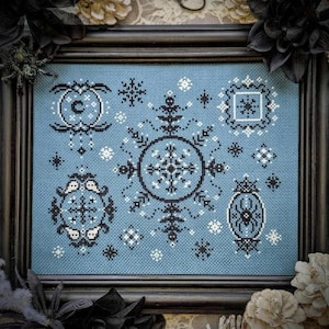 Puede incluir: Arte de punto de cruz enmarcado con un fondo azul claro y patrones geométricos en blanco y negro. El diseño incluye motivos circulares y cuadrados, copos de nieve y una araña. La obra de arte se muestra en un marco negro.