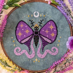 May include: A circular embroidery hoop containing a cross-stitch design on blue fabric. The design features a central moth with purple wings, yellow star accents, and long, curling light purple tails. A white crescent moon sits above the moth.