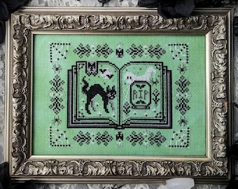 May include: A framed cross-stitch design featuring a green book with a black cat, a ghost, and the word "RIP" on the cover. The book is surrounded by a black and white border with spiders and bats.