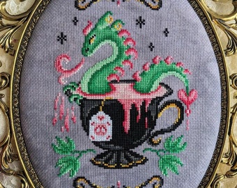 May include: A green dragon with pink accents is curled up inside a black teacup with a pink and white tea bag. The teacup is dripping pink liquid and is surrounded by green leaves and pink hearts.