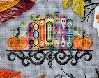 May include: A cross-stitch design featuring a stack of colorful books with intricate patterns, flanked by orange pumpkins. The design is set against a gray background, with fall leaves and embroidery floss spools surrounding the central image.