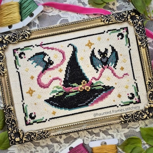 May include: A framed cross-stitch featuring a black witch's hat, bats, and stars. The artwork is in a gold ornate frame, with pink ribbon and spools of thread. The design has a Halloween theme.