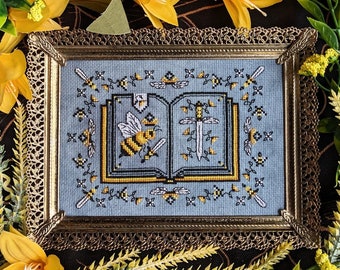 May include: A framed cross-stitch design featuring a book with a bee and a sword on a blue background. The frame is gold with a decorative border.
