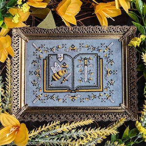 May include: A framed cross-stitch design featuring a book with a bee and a sword on a blue background. The frame is gold with a decorative border.
