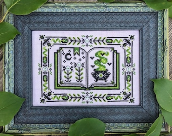 May include: A framed cross-stitch design featuring a book with a cauldron and green smoke. The book is open and the pages are filled with black and white illustrations. The frame is dark green with a gold trim.