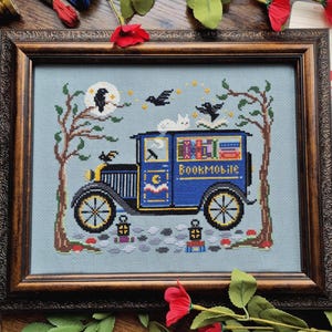 May include: Cross-stitch art in a brown wooden frame. The image depicts a blue "Bookmobile" truck with books on top, a white cat, and black birds. The background is a light blue with trees, a moon, and stars. The text "Bookmobile" is visible on the side of the truck.