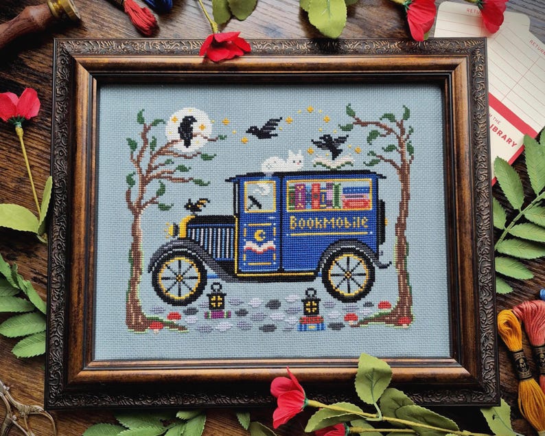 May include: A framed cross-stitch embroidery on light blue fabric featuring a blue vintage truck labeled Bookmobile, carrying colorful books. The scene includes two bare trees, a white cat, black birds, a moon, and two lanterns on a cobblestone path.