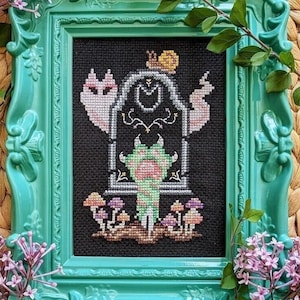 May include: A cross-stitch embroidery framed in a teal green frame. The design features a pink and white cat, a snail, a green monster, and mushrooms.