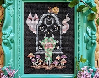 May include: A cross-stitch embroidery framed in a teal green frame. The design features a pink and white cat, a snail, a green monster, and mushrooms.