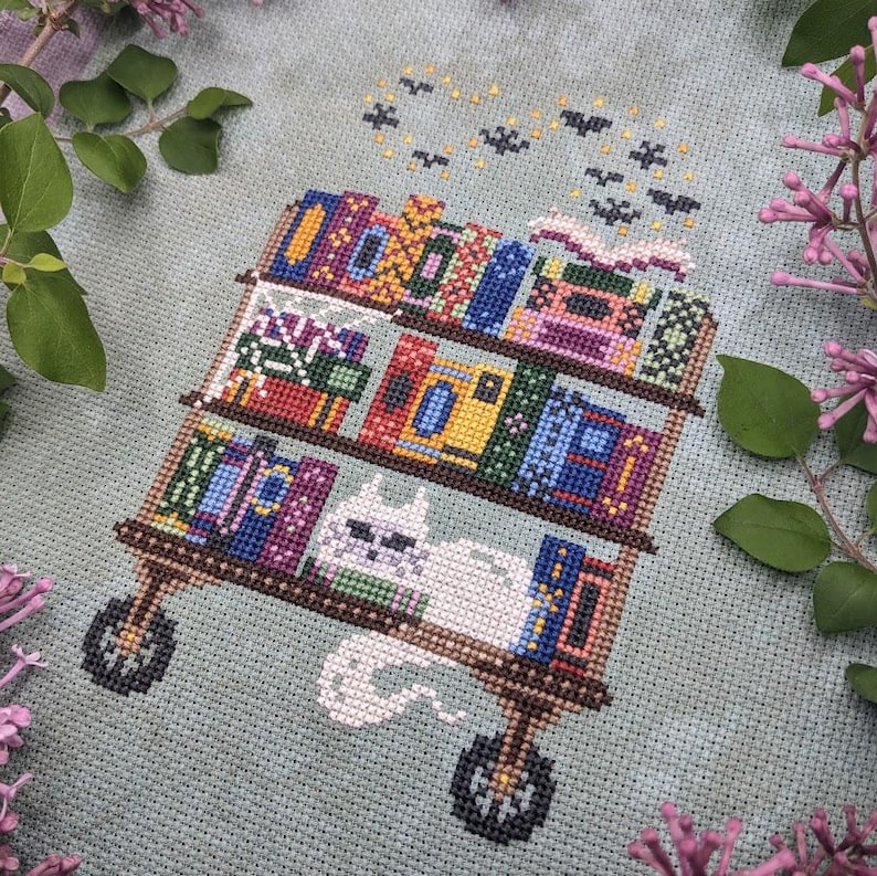 May include: A cross-stitch embroidery on light gray fabric featuring a three-tiered rolling cart filled with colorful books. A white cat rests on the bottom shelf, and a swarm of small black bats flies above the top shelf near an open book.