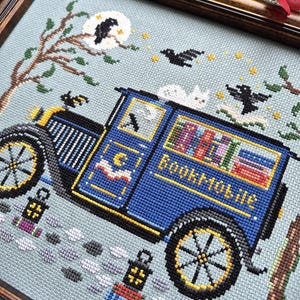 May include: A cross-stitch embroidery in a dark wood frame depicting a blue vintage bookmobile van with the word Bookmobile stitched on the side. The scene includes a white cat on the roof, black birds, a moon, stars, and lanterns on a light blue background.