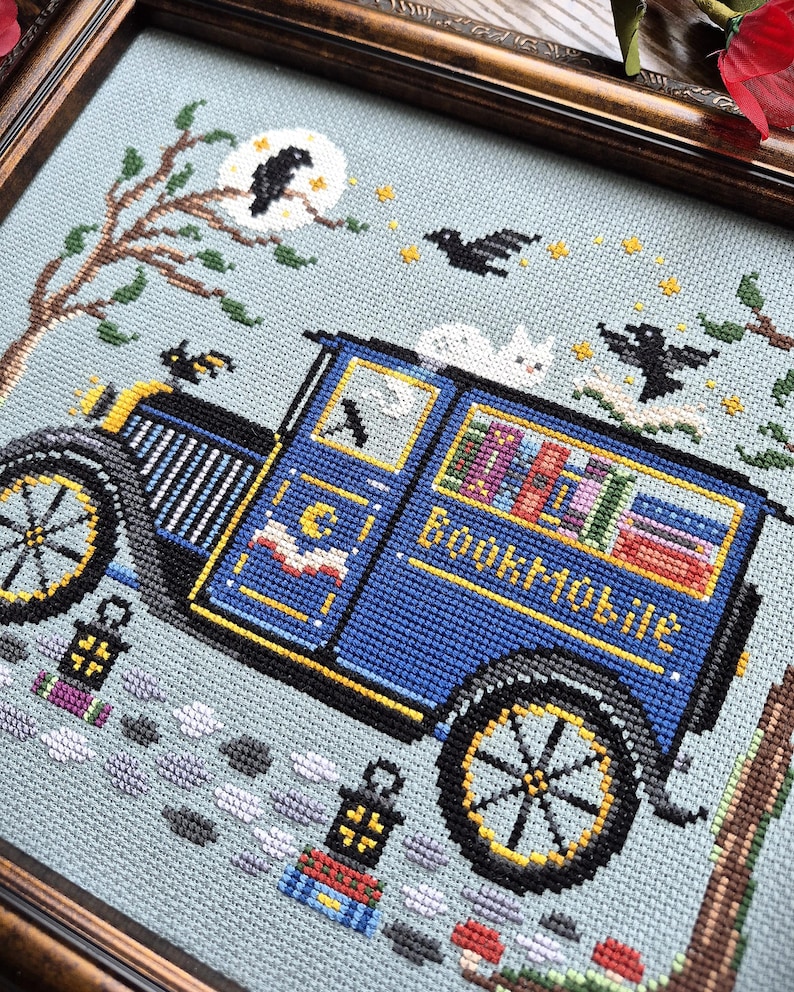 May include: A cross-stitch embroidery in a dark wood frame depicting a blue vintage bookmobile van with the word Bookmobile stitched on the side. The scene includes a white cat on the roof, black birds, a moon, stars, and lanterns on a light blue background.