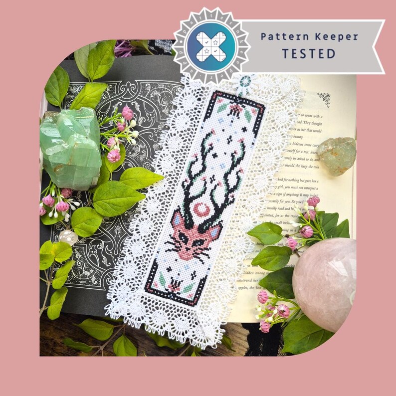 Puede incluir: Un marcap&aacute;ginas rectangular de punto de cruz con una cabeza de gato rosa estilizada con astas negras, rodeado de encaje blanco. El dise&ntilde;o incluye peque&ntilde;os motivos florales y una luna creciente. Una banda superior dice Pattern Keeper TESTED.