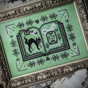 May include: A green cross-stitch embroidery of an open book with a black cat, bats, and a tombstone that reads "RIP". The book is surrounded by a border of black and white leaves and spiders.
