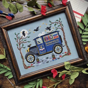 May include: A framed cross-stitch embroidery on light blue fabric featuring a blue vintage van labeled Bookmobile, a white cat on the roof, two black birds, trees, a moon, and stars. The design includes small lanterns and a cobblestone path.