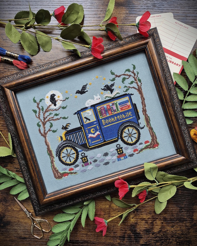 May include: A framed cross-stitch embroidery on light blue fabric featuring a blue vintage van labeled Bookmobile, a white cat on the roof, two black birds, trees, a moon, and stars. The design includes small lanterns and a cobblestone path.