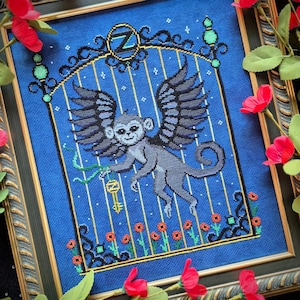 May include: Cross-stitch artwork of a winged monkey holding a key, set within a cage design. The monkey is gray with black wings, set against a blue background. The artwork is framed with a decorative border and red floral accents.