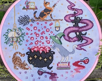 May include: A cross-stitch embroidery hoop with a whimsical design featuring cats, a cauldron, and magical elements. The cats are depicted in various poses, including one wearing a witch's hat and stirring a cauldron. The design is set against a light blue background with pink accents.