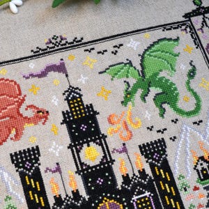 K&ouml;nnte beinhalten: Eine Kreuzstichstickerei auf hellbraunem Stoff mit einer schwarzen Burg mit gelben Fenstern, einem roten Drachen auf der linken Seite und einem gr&uuml;nen Drachen auf der rechten Seite. Das Design enth&auml;lt kleine gelbe und wei&szlig;e Sterne und orange Flammen.