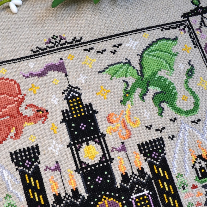 K&ouml;nnte beinhalten: Eine Kreuzstichstickerei auf hellbraunem Stoff mit einer schwarzen Burg mit gelben Fenstern, einem roten Drachen auf der linken Seite und einem gr&uuml;nen Drachen auf der rechten Seite. Das Design enth&auml;lt kleine gelbe und wei&szlig;e Sterne und orange Flammen.