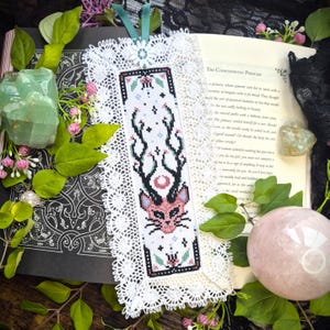 Puede incluir: Un marcap&aacute;ginas rectangular con un dise&ntilde;o de punto de cruz de una cabeza de gato rosa con astas negras, rodeado de peque&ntilde;as estrellas y motivos florales. El bordado est&aacute; enmarcado por un borde de encaje blanco y una cinta de color cerceta.