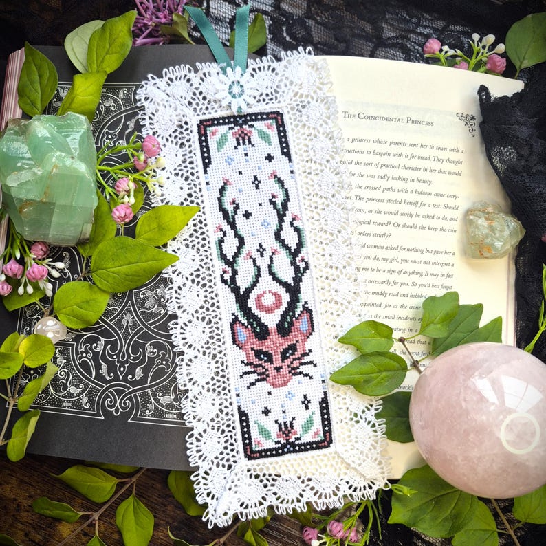 Puede incluir: Un marcap&aacute;ginas rectangular con un dise&ntilde;o de punto de cruz de una cabeza de gato rosa con astas negras, rodeado de peque&ntilde;as estrellas y motivos florales. El bordado est&aacute; enmarcado por un borde de encaje blanco y una cinta de color cerceta.