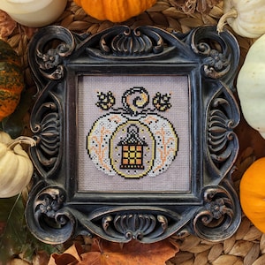 May include: A framed cross-stitch design featuring a white pumpkin with a lantern inside. The pumpkin is surrounded by orange and yellow leaves and vines. The frame is black with ornate details.