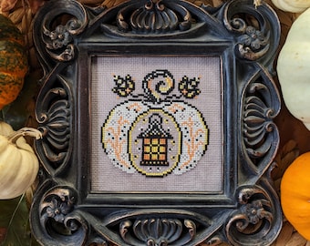 May include: A framed cross-stitch design featuring a white pumpkin with a lantern inside. The pumpkin is surrounded by orange and yellow leaves and vines. The frame is black with ornate details.