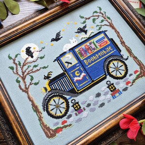May include: A cross-stitch embroidery in a dark wood frame depicting a blue bookmobile van with the text Bookmobile on the side. The scene includes a tree, a moon with a black bird, flying birds, a white cat, and two lanterns on a light blue background.