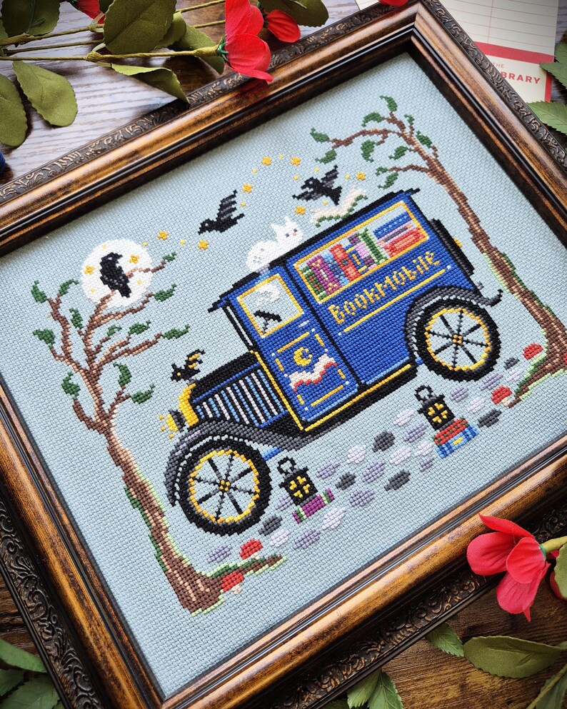 May include: A cross-stitch embroidery in a dark wood frame depicting a blue bookmobile van with the text Bookmobile on the side. The scene includes a tree, a moon with a black bird, flying birds, a white cat, and two lanterns on a light blue background.