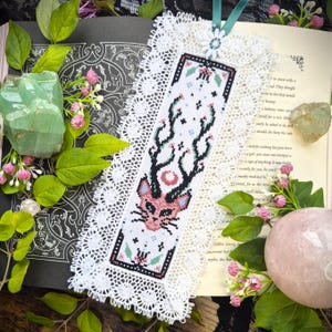 May include: A handmade bookmark with a cross-stitch design featuring a pink and black deer-like creature with antlers. The bookmark is white with a decorative lace edge and a teal ribbon. The bookmark is placed on an open book.