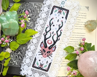 May include: A handmade bookmark with a cross-stitch design featuring a pink and black deer-like creature with antlers. The bookmark is white with a decorative lace edge and a teal ribbon. The bookmark is placed on an open book.