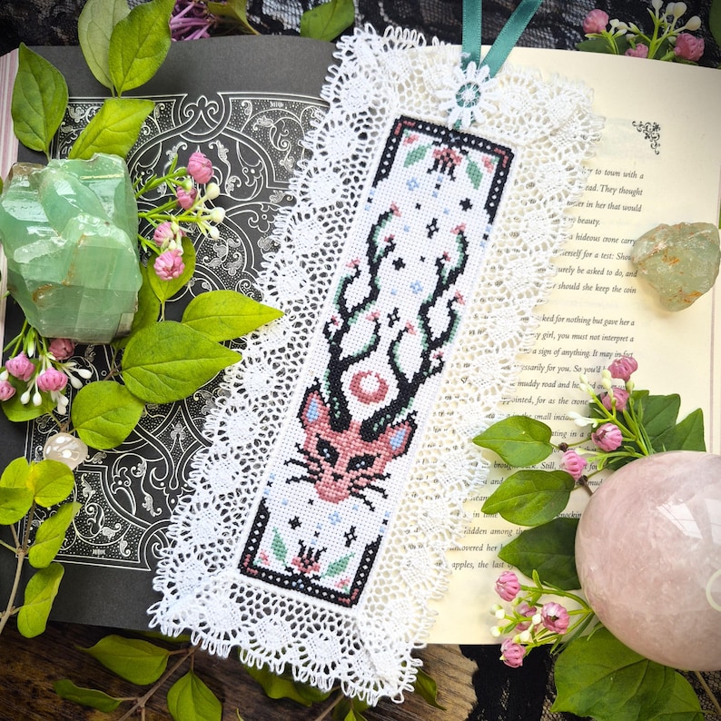 Puede incluir: Un marcap&aacute;ginas rectangular con un dise&ntilde;o de punto de cruz de una cabeza de gato rosa con grandes astas, rodeado de peque&ntilde;as flores y estrellas, enmarcado por un borde de encaje blanco y rematado con una cinta azul verdosa.