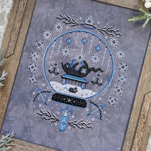 May include: A cross-stitch pattern of a snow globe with two black cats inside, sitting on a small table. The snow globe is surrounded by snowflakes and other wintery designs.