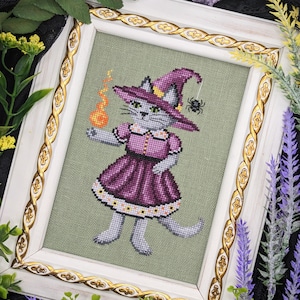 Peut inclure: Un motif de point de croix représentant un chat gris habillé en sorcière, portant une robe violette et un chapeau pointu, tenant un orbe orange brillant. Le chat est debout sur un fond vert avec une araignée au-dessus.