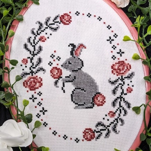 May include: A gray bunny rabbit cross-stitch embroidery design with pink flowers and leaves in a white hoop.