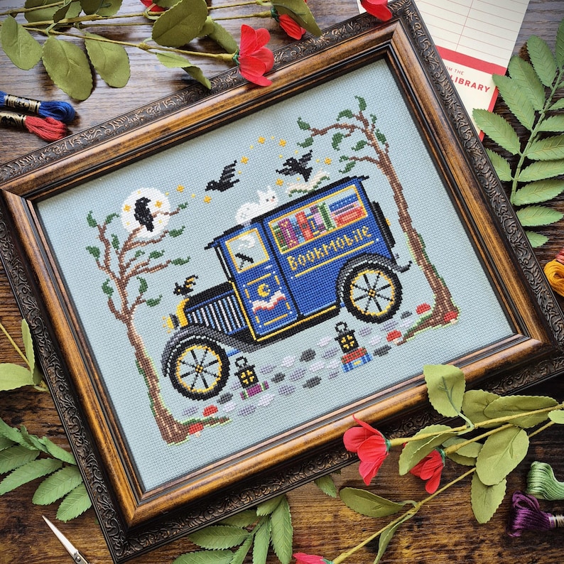 May include: A framed cross-stitch embroidery on light blue fabric featuring a blue vintage van labeled Bookmobile, carrying books. The scene includes a white cat on the roof, two black birds, a full moon, trees, and two small lanterns.