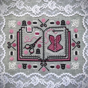 May include: A cross-stitch embroidery on gray fabric featuring an open book with sewing motifs including a corset, scissors, thread spools, and thimbles in pink, black, and white. The design is framed by a white floral lace border.