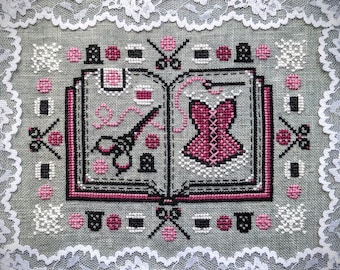 May include: A cross-stitch embroidery design featuring a book with a pink corset and scissors on the left page and a pink corset on the right page. The design is surrounded by a white lace border.